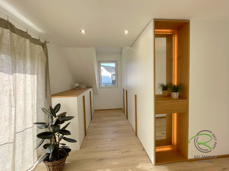 Maßgefertigter Einbauschrank mit Dachschräge in Tuttlingen von Schreinerei Holzdesign Ralf Rapp in weiß und Eiche mit halbhoher Kommode und raumhohen Dachnischenschrank und auf der Stirnseite Anrichte mit Spiegel und indirekter Beleuchtung