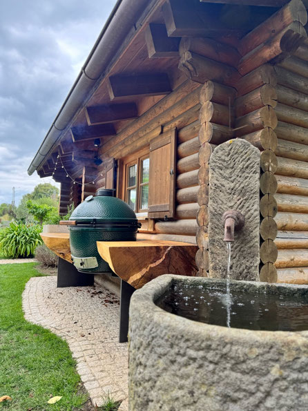Big Green Egg in massiver Geisinger Esche mit naturgeschwungener Baumkante vor alter Blockhütte und Natursteinbrunnen bei Geisingen