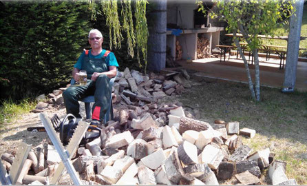 Michael Kothe in Schutzkleidung und mit Kettensäge inmitten eines Haufens von Brennholz. er trinkt Bier