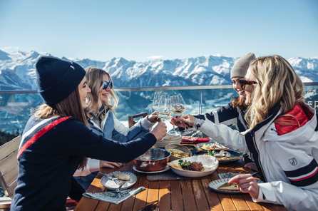 Vier Personen stoßen beim Essen auf einer Terrasse mit Blick auf das verschneite Alpenpanorama an.