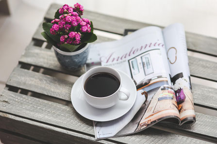 Auf einer Holzkiste liegen zwei gefaltete Zeitungen. Darauf steht eine weiße Tasse mit Kaffee auf einer Untertasse. Schräg hinter der Tasse ein Blumentopf mit einer pink blühenden Pflanze.