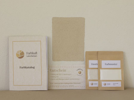 Farbhaft-Farbkatalog, Farbmuster und Gutschein stehen vor einer hellen Lehmwand mit gestrichener Testfläche zur Prüfung von Farbton, Oberfläche und Untergrund.
