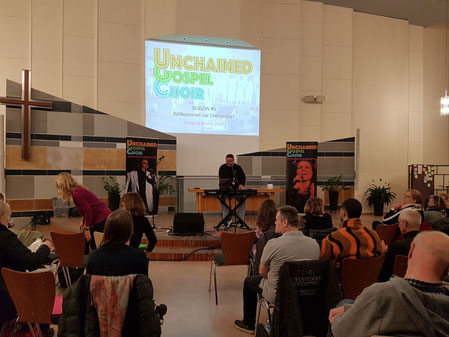 Kirchensaal in der EFG Tempelhof: Sicht mittig von hinten auf die Bühne, rechts und links sitzen Chorsänger:innen, Hanjo sitzt auf der Bühne am Klavier, an die Wand hinter ihm ist eine Willkommensnachricht an den Chor projiziert