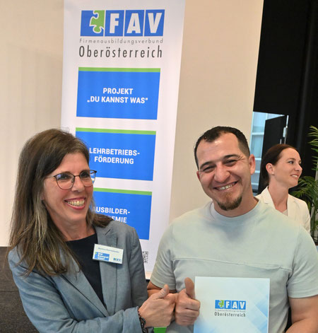 Martina Reichetzeder und ein Teilnehmer stehen lächelnd vor einem FAV OÖ-Roll-Up und halten gemeinsam ein Zertifikat in der Hand – symbolischer Moment der Anerkennung.