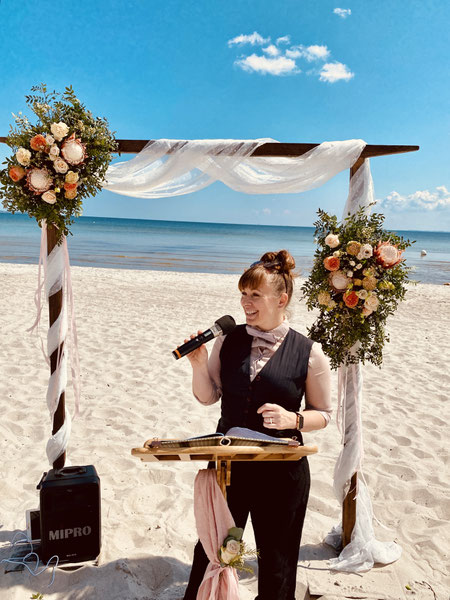 Constanze Marienfeld spricht bei einer Trauung am Strand ins Mikro, sie steht vor einem mit Blumen geschmückten Traubogen