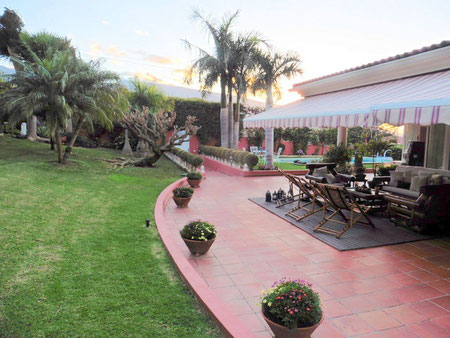 Blick vom subtropischen Garten auf die Terrasse mit Holzmöbel, dem Haus und dem Pool von der Immobilie.