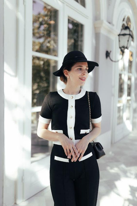 Elegant woman in a black and white outfit with a hat and gold details in front of a bright building facade.
