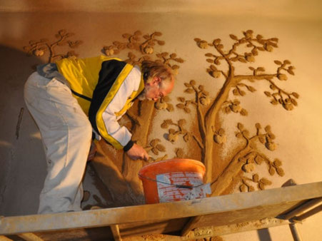 Person bei der handwerklichen Gestaltung eines Apfelbaum-Motivs auf einer Wandfläche mit Lehmfarbe.