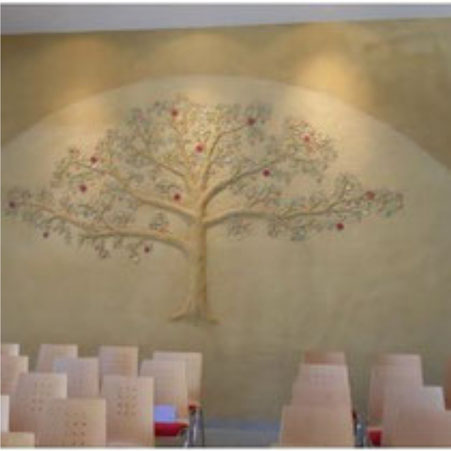 Plastisch aus Lehmputz gestaltetes und mit Lehmfarbe bemaltes Baum-Motiv an der Wand eines modernen Kirchensaals.