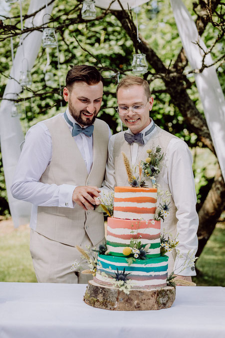 zwei Männer schneiden gemeinsam eine bunte Hochzeitstorte an bei gleichgeschlechtlicher Hochzeitsfeier im Saarland