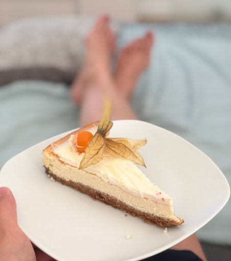 American Cheese Cake auf einem weißen Teller. Frau auf dem Sofa hält den Teller mit Käsekuchen und einer Physalis in der Hand.