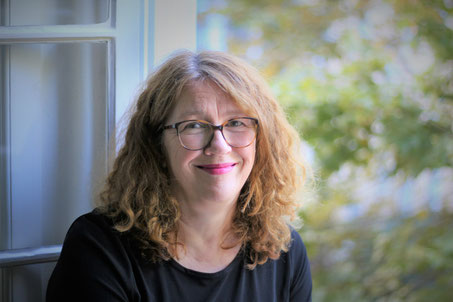 Trauerrednerin Margit Vida sitzt vor einem geöffneten Fenster und lächelt. Sie hat braune Haare, trägt eine Brille und ein schwarzes Kleid.