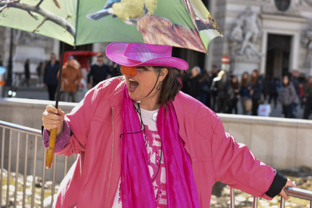 Susanne Christine Drdla mit schillerndem pinken Cowboyhut, Regenschirm mit großen Vögeln bei strahlendem Sonnenschein, und einer roten Clownsnase