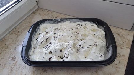 Eine schwarze Kunststoffschale gefüllt mit einer großen Portion Stracciatella-Eis oder Vanilleeis mit Schokoladensauce auf einer hellen Marmor-Fensterbank.