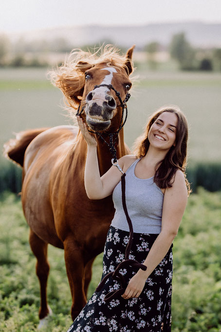 Ein braunes Pferd schüttelt den Kopf während ein Mädchen daneben steht und darüber lächelt