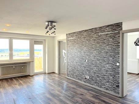 Helles Wohnzimmer in Neuötting mit grauer Steinwand-Optik, modernem Laminatboden, großem Fenster und Zugang zum sonnigen Balkon mit herrlichem Ausblick über die Umgebung von Neuötting im Landkreis Altötting.