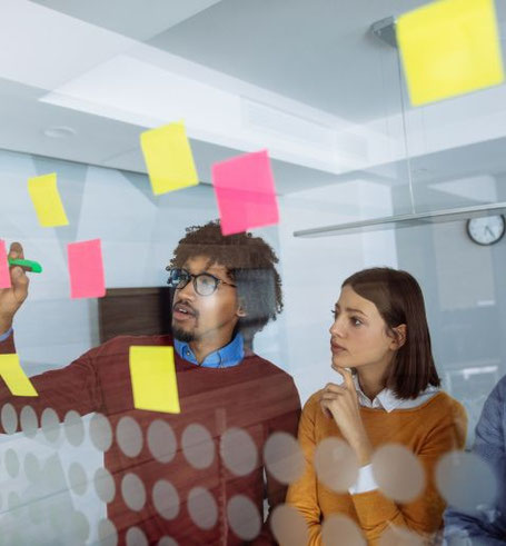 Zwei Personen stehen vor einem durchsichtigen Whiteboard. Auf dem Whiteboard sind Post-it-Zettel geklebt, eine Person schreib mit einem Marker auf das Board.