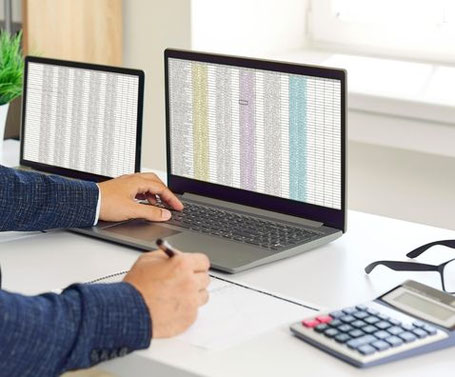 Eine Person sitzt am Schreibtisch. Zwei Laptops sind geöffnet, auf deinen ist Excel geöffnet.