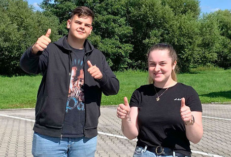 Ein junger Mann und eine junge Frau, beide mit schwarzem Outfit und blauen Jeans heben beide Hände mit dem „Daumen nach oben“-Zeichen.