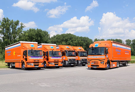 Ein Elektro LKW steht quer geparkt vor weiteren vier Elektro LKW. Alle sind in orange und mit dem Logo der Spedition versehen.