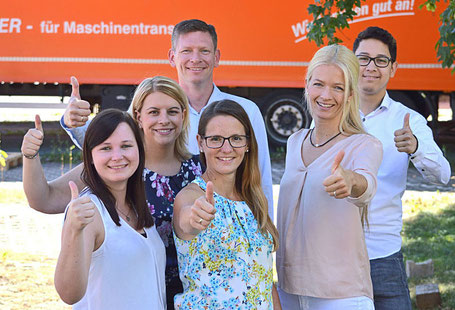 Sechs Mitarbeiter in unterschiedlicher Kleidung machen alle das „Daumen hoch“-Zeichen. Im Hintergrund steht ein Lkw-Anhänger.