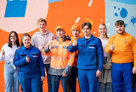 8 Azubis stehen, zum Teil mit typischer orangener Rüdinger Kleidung vor dem Verwaltungsgebäude der Rüdinger Spedition Die Plane ist bunt bedruckt. Alle lachen oder schauen fröhlich.