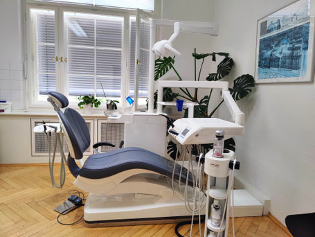 Behandlungszimmer der Zahnarztpraxis Doumit in Berlin-Friedenau mit moderner Behandlungseinheit, grauem Polster, Airflow Prophylaxis Master und Pflanze im Hintergrund.