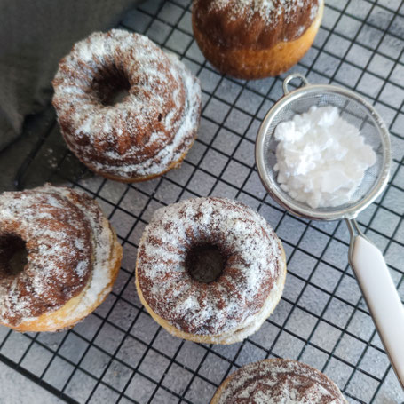 Mehrere mit Puderzucker bestäubte Mini-Gugelhupfe auf einem Kuchengitter, daneben ein Sieb mit Puderzucker