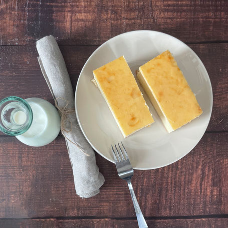 Zwei Stück Rahm-Käsekuchen auf einem Teller mit Gabel und Stoffserviette und einer Karaffe mit Milch daneben