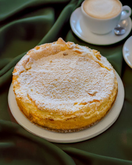 Eine mit Puderzucker bestäubte Käsetorte auf einem Kuchenteller, dahinter eine Tasse Cappuccino