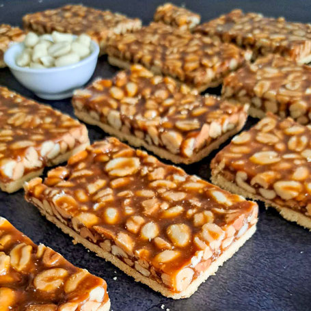 Mehrere rechteckige Stücke des Gebäcks „Salzige Nuss trifft Butterkaramell“ liegen in Reihen auf dunkler Oberfläche. In der Mitte steht eine weiße Schale mit geschälten Erdnüssen.
