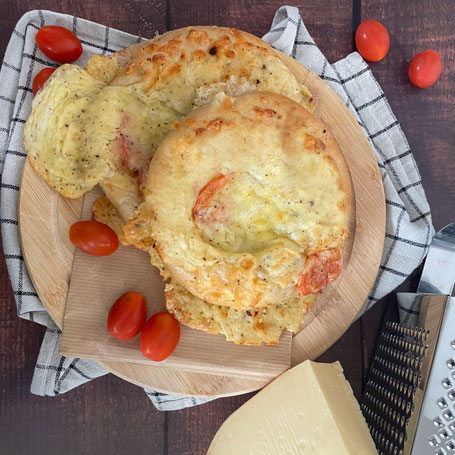 Zwei mit Käse überbackene Tomaten-Käse-Brötchen liegen übereinander auf einem runden Holzbrett, daneben liegen frische Tomate sowie ein Gouda mit einer Käsereibe