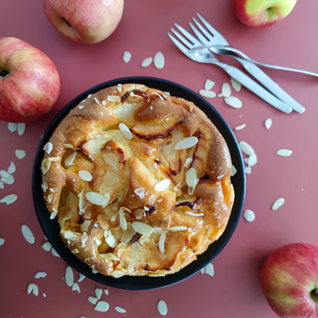 Eine Apfeltorte steht auf einem schwarzen Teller., daneben liegen Äpfel und ein paar Gabeln