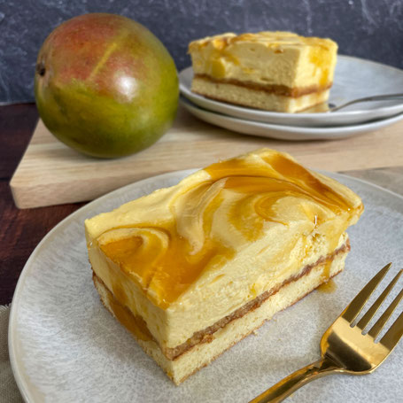 Ein Stück Mango-Sahne-Kuchen mit cremiger Schicht und Mangoswirl auf einem grauen Teller, daneben eine Gabel und im Hintergrund eine ganze Mango und ein weiteres Kuchenstück auf Tellern