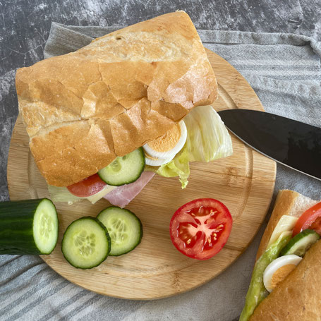 Ein mit Kochschinken, Gouda, Salat, Gurke, Tomate und Ei belegtes Baguette auf einem Holzbrett, daneben aufgeschnitte Gurke und Tomate