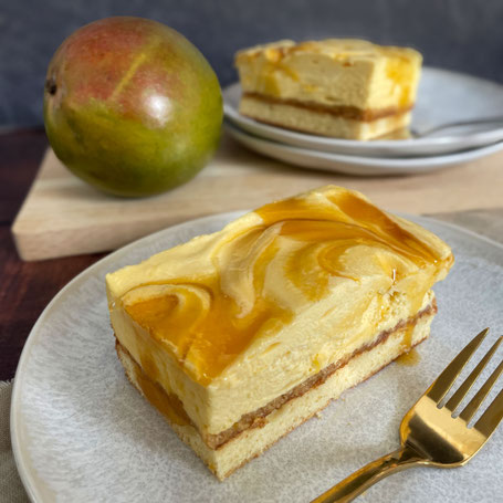 Ein Stück Mango-Sahne-Kuchen mit cremiger Schicht und Mangoswirl auf einem grauen Teller, daneben eine Gabel und im Hintergrund eine ganze Mango und zwei weitere Kuchenstücke auf Tellern