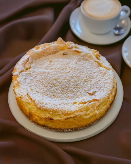 Eine mit Puderzucker bestäubte Käse-Mohntorte auf einem Kuchenteller, dahinter eine Tasse Cappuccino