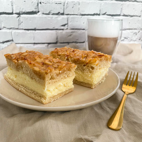 Zwei Stücke gefüllter Bienenstich mit Vanillecreme auf einem Teller, daneben eine goldene Gabel und ein Glas Latte Macchiato