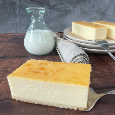 Ein Stück Rahm-Käsekuchen auf Kuchenheber, im Hintergrund weitere Stücke auf Tellern und eine Karaffe mit Milch