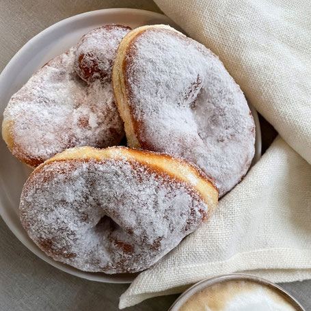 Drei zuckrige Krapfen auf einem weißen Teller, im Anschnitt ein helles Tuch und eine Tasse Cappuccino