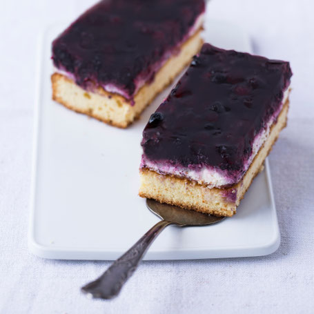 Zwei Stücke Heidelbeerkuchen auf einem Teller, daneben ein Messer und ein paar Heidelbeeren