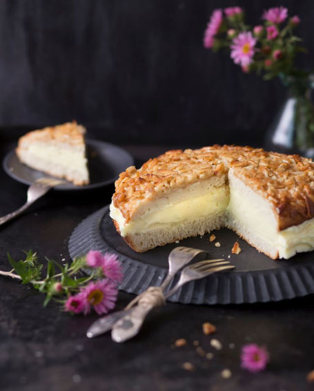 Eine angeschnittene Bienenstichtorte auf einem dunklen Blech, im Hintergrund ein Telle rmit enem Stück Bienenstichtorte