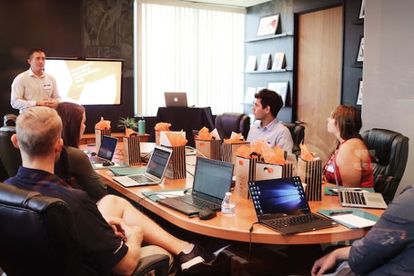 Fünf Personen sitzen um einen Besprechungstisch und diskutieren ein Projekt. Ein Projektleiter steht vor einem Bildschirm und leitet das Meeting. Kommunikation für den nächsten Schritt. Foto: Campaign Creators (Unsplash)