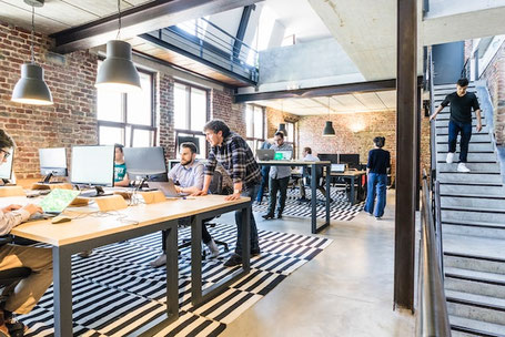 Büroarbeit in modernem, fabrikähnlichem Raum. Zwei Männer kommunizieren an einem Pult. Eine Person kommt die Treppe herunter. Strategischer Aufbau von Produktkommunikation. Foto: Proxyclick Visitor Management System (Unsplash)