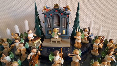 Ein Engels-Orchester aus kleinen Holzfiguren gibt ein Konzert. Im Hintergrund eine historische blaue Holzorgel, an der ein Engelchen sitzt.