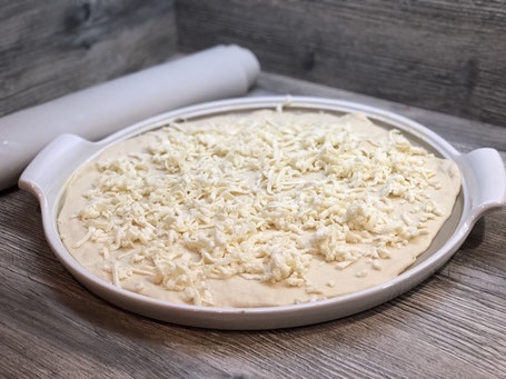Runde weißer Stoneware-Form auf Arbeitspalatte mit Fett bestrichen, Pizzateig belegt und Käse bestreut