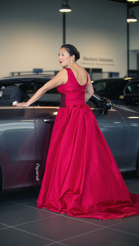 Robe de mariée rouge recouverte d'une large dentelle uniquement sur le devant au niveau de la taille, jupe à plis plats terminée par une longue traîne