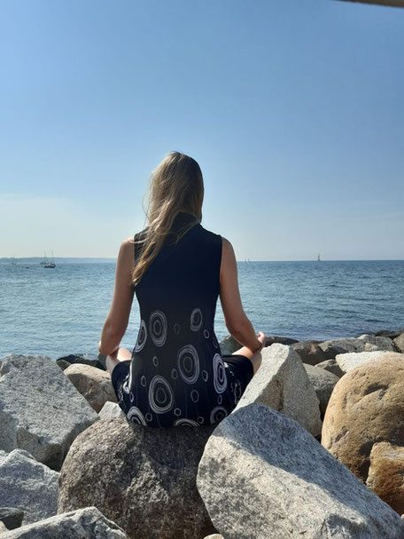 Ramona sitzt mit Rücken zur Kamera auf großen Steinen am Ufer und schaut im Schneidersitz auf das Meer.