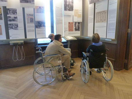 Drei Teilnehmer einer Fortbildung erkunden eine Ausstellung in der für sie ungewohnten Weise aus dem Rollstuhl heraus.