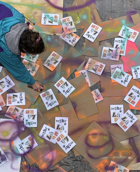 Das Bild ist von oben aufgenommen. Auf dem Boden liegen viele Pappkertons und Zines, auf denen mehr*Lärm steht. Eine Person sprüht mit einer Sprühdose Farbe über die Kartons und Zines.